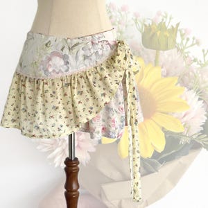 May include: A wrap skirt with a floral pattern in cream, yellow, and pastel colours. The skirt features a ruffled hem and a tie closure. The fabric has a mix of floral and small dot patterns, creating a vintage aesthetic.