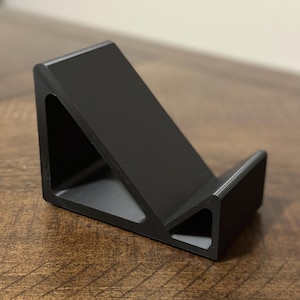 May include: A black, angled phone stand. The stand is made of a solid material and features a triangular cutout on the side. The stand is designed to hold a phone or tablet at an angle for easy viewing.