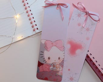 May include: Two pink and white bookmarks with pink ribbon ties. One bookmark features a Hello Kitty illustration holding a mug. The other bookmark has a pink heart and star design. A spiral notebook and fairy lights are in the background.
