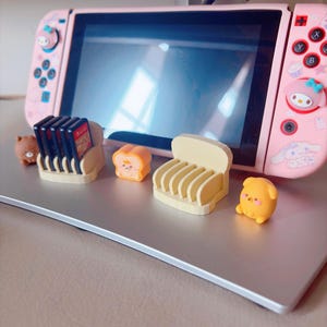 May include: A pink Nintendo Switch console with decorative stickers and a collection of game cartridges. The console is accompanied by several small, cute figurines and game cartridge holders, including a bear, toast, and a chair.