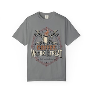 May include: A grey short-sleeve t-shirt featuring a graphic of a coffee mug with steam, crossed wrenches, and gears. The text reads COFFEE. WORK. REPEAT. and Built for the Grind. The design is enclosed in a circular frame with red and blue accents.