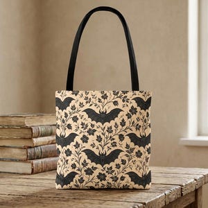 Può includere: Una borsa tote con manici neri caratterizzata da un motivo ripetuto di pipistrelli neri e viticci floreali scuri su uno sfondo beige chiaro. La borsa ha una forma rettangolare ed è realizzata in tessuto con una stampa uniforme sul davanti.