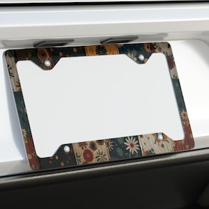 May include: A decorative license plate frame with a floral pattern in shades of brown, red, and green. The frame is mounted on the back of a white car. The design features various flowers and leaves.