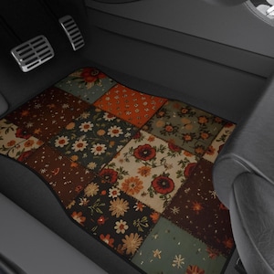 May include: A rectangular car floor mat featuring a patchwork design of various floral patterns in shades of dark brown, orange, cream, and muted green. The mat is positioned on the floor of a vehicle interior with black leather seats and pedals.