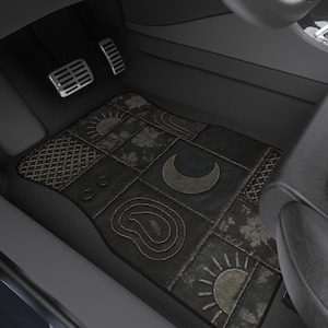 Puede incluir: Una alfombrilla de coche rectangular con un diseño de patchwork gris oscuro que incluye varios patrones como una luna creciente, soles, motivos florales y texturas geométricas. La alfombrilla está colocada en el suelo de un vehículo con asientos negros.