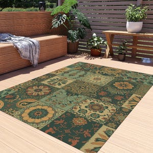 Puede incluir: Alfombra rectangular para exteriores con diseño de patchwork. La alfombra presenta varios patrones florales y paisley en tonos verdes, dorados y óxido. La alfombra está colocada sobre una terraza de madera, con un banco y plantas al fondo. Mide aprox. 200cm x 300cm.