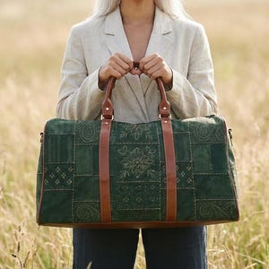 以下が含まれることがあります： 茶色の革のハンドルとアクセントが付いた、緑色のパッチワークのダッフルバッグ。バッグには刺繍された花の模様があり、明るい色のブレザーを着た人が持っており、背の高い草の野原を背景にしています。