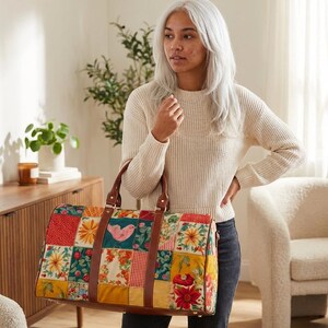 May include: A rectangular duffel bag featuring a colorful patchwork design with floral and bird patterns in shades of red, yellow, and teal. The bag includes brown leather handles and trim, with gold-toned metal hardware accents on the sides.