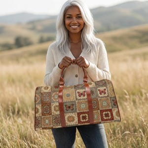 May include: A rectangular duffel bag featuring a patchwork pattern of squares in shades of brown, tan, and cream, decorated with butterfly motifs. The bag includes two brown leather handles and a matching leather trim along the front panel.