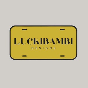 K&ouml;nnte beinhalten: Ein rechteckiges senfgelbes Nummernschild mit abgerundeten Ecken und einem dicken schwarzen Rand. Der Text LUCKIBAMBI ist in einer fetten schwarzen Serifenschrift gedruckt, darunter zentriert das Wort DESIGNS in einer kleineren serifenlosen Schrift.