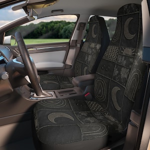Puede incluir: Fundas de asiento de coche negras con un diseño de patchwork con lunas crecientes, soles y patrones geométricos. Las fundas están en el interior de un coche con volante y vista a un campo verde.