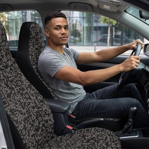 May include: Car seat covers with a repeating black and gray pixelated pattern. The covers are installed in a car, with the driver visible behind the wheel. The car's interior is visible, including the dashboard and steering wheel.