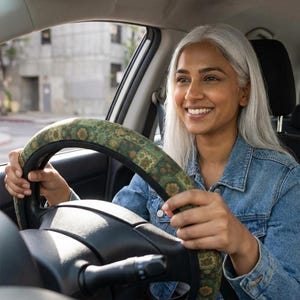May include: A steering wheel cover featuring a dark green fabric with a repeating floral and geometric pattern in shades of gold, brown, and muted orange. The cover is fitted over a black steering wheel, showing a textured surface and fabric.