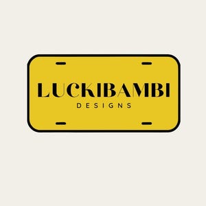 May include: A rectangular yellow license plate with rounded corners and a black border. The text LUCKIBAMBI is printed in a bold black serif font, with the word DESIGNS in a smaller black sans-serif font underneath. Four mounting slots are visible.