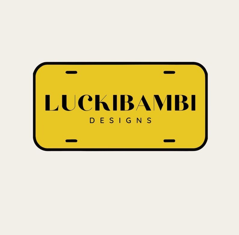May include: A rectangular yellow license plate with rounded corners and a black border. The text LUCKIBAMBI is printed in a bold black serif font, with the word DESIGNS in a smaller black sans-serif font underneath. Four mounting slots are visible.