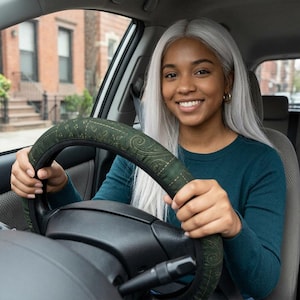 May include: A dark green steering wheel cover featuring a subtle, repeating gold paisley pattern. The cover is fitted over a black steering wheel, providing a textured grip with a decorative design that wraps around the circular rim of the wheel.