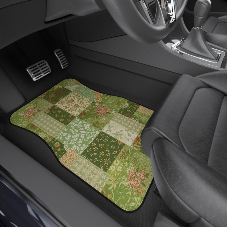 May include: A rectangular car floor mat featuring a patchwork design of various green, olive, and cream floral and botanical patterns. The mat is positioned in the driver side footwell of a vehicle with black interior trim and metal pedals.