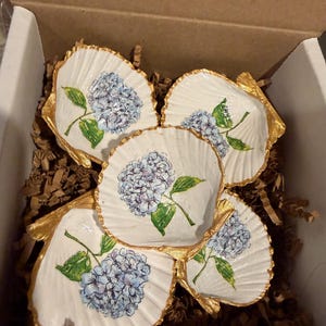 Puede incluir: Un conjunto de cuatro conchas de vieira blancas, cada una con una flor de hortensia azul pintada a mano con hojas verdes. Los bordes de cada concha están acabados con un borde dorado metálico y dispuestas dentro de una caja.