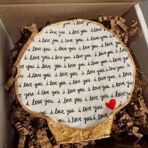 以下が含まれることがあります： 「I love you」という言葉が黒い筆記体で繰り返し書かれた、ハート型の白いオーナメント。小さな赤いハートが見えます。オーナメントは金色の縁取りがあり、茶色の梱包材の中にあります。