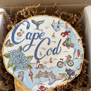Könnte beinhalten: Runde Holzplakette mit einer Karte von Cape Cod, die eine Walflosse, Leuchttürme, ein Fahrrad und den Text "Cape Cod" in blauer Schrift zeigt. Die Plakette hat einen goldfarbenen Rand und befindet sich in einem Karton mit braunem Papier.