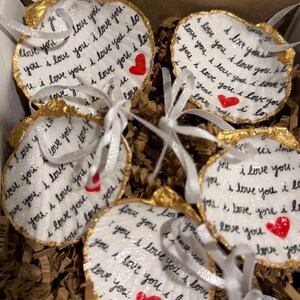 Peut inclure: Cinq ornements en coquillage blanc avec des bords dorés et des rubans blancs. Chaque coquillage porte l'inscription manuscrite "I love you" et un petit cœur rouge. Les ornements sont présentés dans une boîte en carton avec du papier brun.