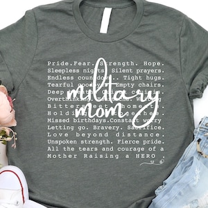 May include: A gray t-shirt with the words "military mom" in white script. The shirt is surrounded by a pair of white sneakers, distressed blue jeans, and a bouquet of pink flowers. The shirt's text includes phrases like "Pride. Fear. Strength. Hope."