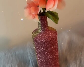 May include: A pink glitter vase with a gold glitter stripe, holding a bouquet of peach-colored artificial flowers.