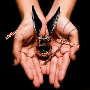 Peut inclure: Un collier plaqué or avec un pendentif en forme de snitch doré et un charme de baguette. Le snitch est ouvert et a des ailes noires. Le texte "18K Gold-plated" est visible sur l'image.