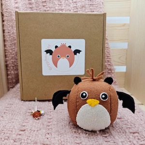 May include: A brown plush owl with black wings, a white belly, and a yellow beak sits on a pink surface. A small, detailed needlework piece and a cardboard box with a sticker featuring a cartoon character are also present.
