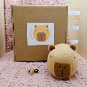 May include: A tan capybara plush toy with a stitched face and small ears sits next to a brown cardboard box with a matching capybara sticker. A small needle and thread are also visible.