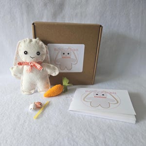 May include: A white felt bunny rabbit sewing kit with a red and white checkered ribbon, a carrot, and a needle and thread. The kit comes in a brown cardboard box with a label that says "Make Your Own Bunny Rabbit".