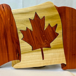 May include: Wooden sculpture of the Canadian flag. The flag has a maple leaf design in the centre, with red and light-coloured wood sections. The flag is designed to appear as if it is waving.