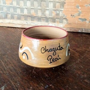 May include: A small, brown ceramic cup with a red rim and white and black painted designs. The cup says "Chazuta Peru" in black lettering.
