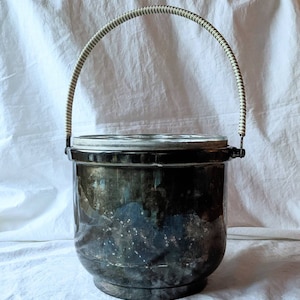 May include: A silver ice bucket with a white and black striped handle. The bucket has a glass lid and a slightly rounded shape.
