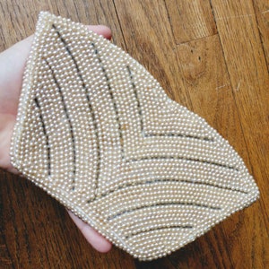 Puede incluir: Un bolso de mano blanco de cuentas con un patrón geométrico. El bolso está hecho de pequeñas cuentas redondas que están cosidas juntas en un patrón repetitivo.