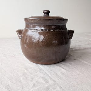 May include: A brown ceramic pot with a lid, featuring two side handles. The pot has a rounded shape and a textured surface. The lid has a small knob on top. The pot is sitting on a white surface.