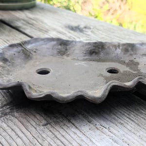 Puede incluir: Un platillo para plantas de hormigón gris con borde ondulado y dos orificios de drenaje. Descansa sobre una superficie de madera envejecida. El diseño es sencillo y rústico, adecuado para plantas pequeñas o suculentas.