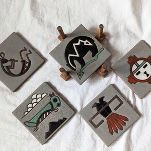 May include: Set of five gray ceramic coasters with Native American-inspired designs. The coasters feature a flute player, a bear, a Kachina doll, a grasshopper, and a bird. The coasters are displayed on a wooden stand.