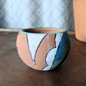 Może przedstawiać: Mała, okrągła doniczka z terakoty z ręcznie malowanym wzorem. Doniczka ma abstrakcyjne kształty w kolorze białym, niebieskim, zielonym i czarnym na jasnobrązowym tle.
