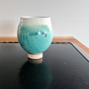 May include: A handmade ceramic vessel with a rounded body and a short, cylindrical base. The upper portion is white, transitioning to a vibrant turquoise hue. The vessel sits on a dark surface, showcasing its unique shape and color.