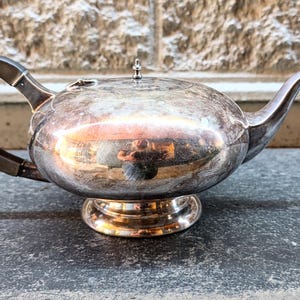May include: A silver-colored teapot with a dark handle and spout. The teapot has a rounded body and sits on a decorative base. The handle is dark brown. The teapot is likely made of metal and has a reflective surface.