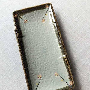 May include: A rectangular ceramic plate with a light blue glaze and a brown rim. The plate has a textured surface and is decorated with small yellow and red flowers.