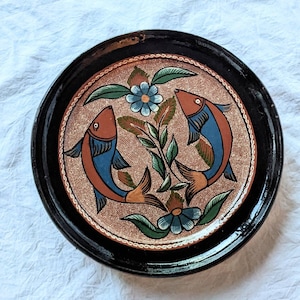 May include: A round ceramic plate with a black rim and a brown background. The plate is decorated with two blue and orange fish facing each other, surrounded by green leaves and blue flowers.