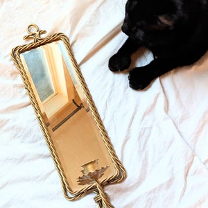 Peut inclure: Un miroir en laiton vintage de forme rectangulaire avec un cadre torsadé en forme de corde. Le miroir a un dessus décoratif et un bougeoir en bas. Un chat noir est couché sur un drap blanc à côté du miroir.