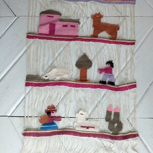 May include: A woven wall hanging with a colorful design featuring a house, a tree, a llama, a sheep, and two figures. The hanging is made of white yarn with red, pink, brown, green, and blue accents.