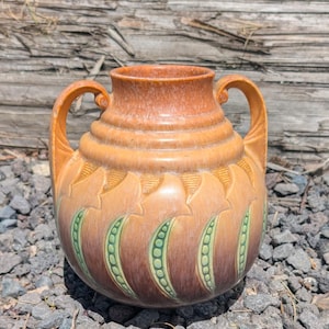 Puede incluir: Un jarrón de cerámica con dos asas, en tonos naranja, marrón y verde. El jarrón tiene una superficie texturizada con un diseño repetido de hojas y vainas, adecuado para la decoración del hogar.