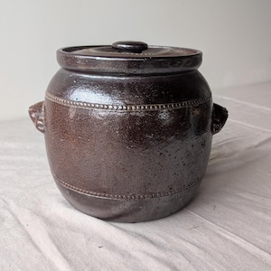 May include: A dark brown ceramic jar with a lid. The jar has two small handles on the sides and a textured surface. The lid has a small knob on top. The jar is sitting on a white cloth.