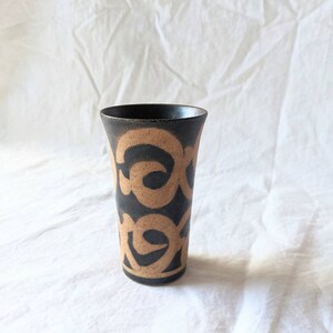May include: A black ceramic vase with a brown abstract pattern. The vase has a cylindrical shape and a slightly flared rim.