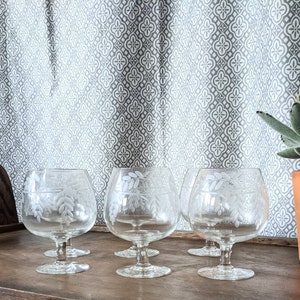 Może przedstawiać: Zestaw sześciu przezroczystych szklanek do brandy z delikatnym, grawerowanym wzorem kwiatowym. Szklanki są ułożone na drewnianej powierzchni.