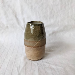 Puede incluir: Un jarrón pequeño de cerámica con un esmalte marrón claro y verde. El jarrón tiene un cuerpo redondeado y una abertura ligeramente acampanada.